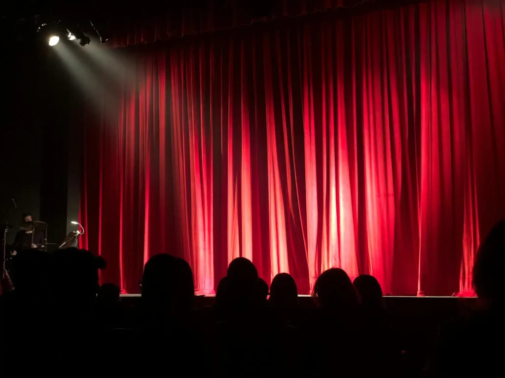 Nell'immagine si vede il palco di un teatro con un sipario rosso e la gente seduta in platea.